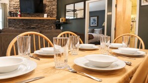 A dining table set for six with white plates, clear glasses, and cutlery, in a room with a stone wall, a mounted TV, and a cross on the wall. - Bearmont- Pool, HotTub, FirePit, AC, Gym, Sauna (Killington)