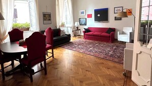 Un salon spacieux doté d’un parquet en bois disposé en point de Hongrie, d’un canapé rouge, d’une table à manger avec des chaises et d’un ventilateur de plafond.