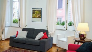 Un salon avec un canapé gris, un fauteuil rouge, une petite table blanche avec une lampe et un tableau encadré au mur.