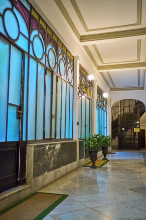 A spacious corridor with ornate stained glass windows, a marble floor, and potted plants.