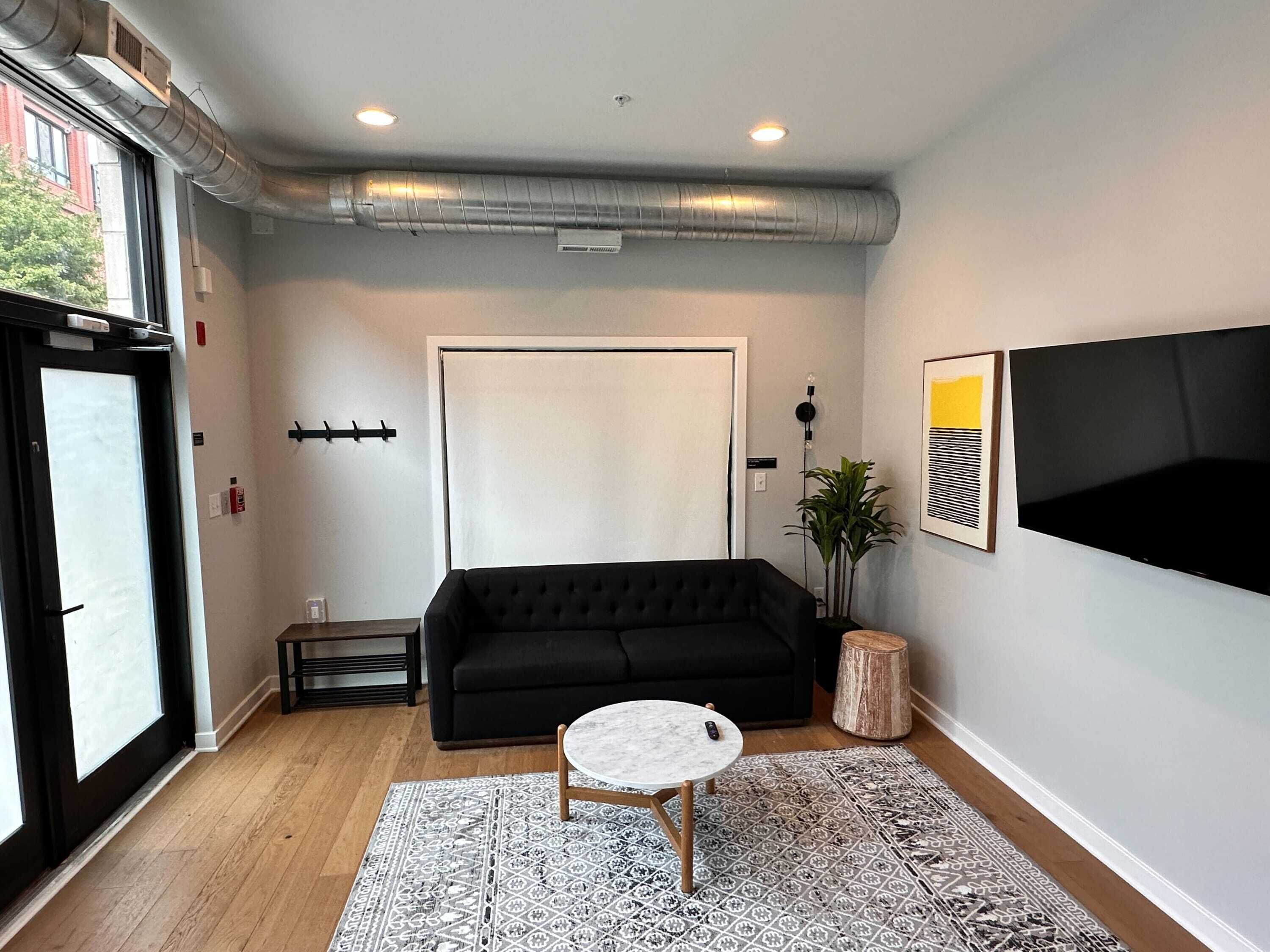 A modern living room with a black sofa, a small round table, a patterned rug, and a large TV on the wall.
