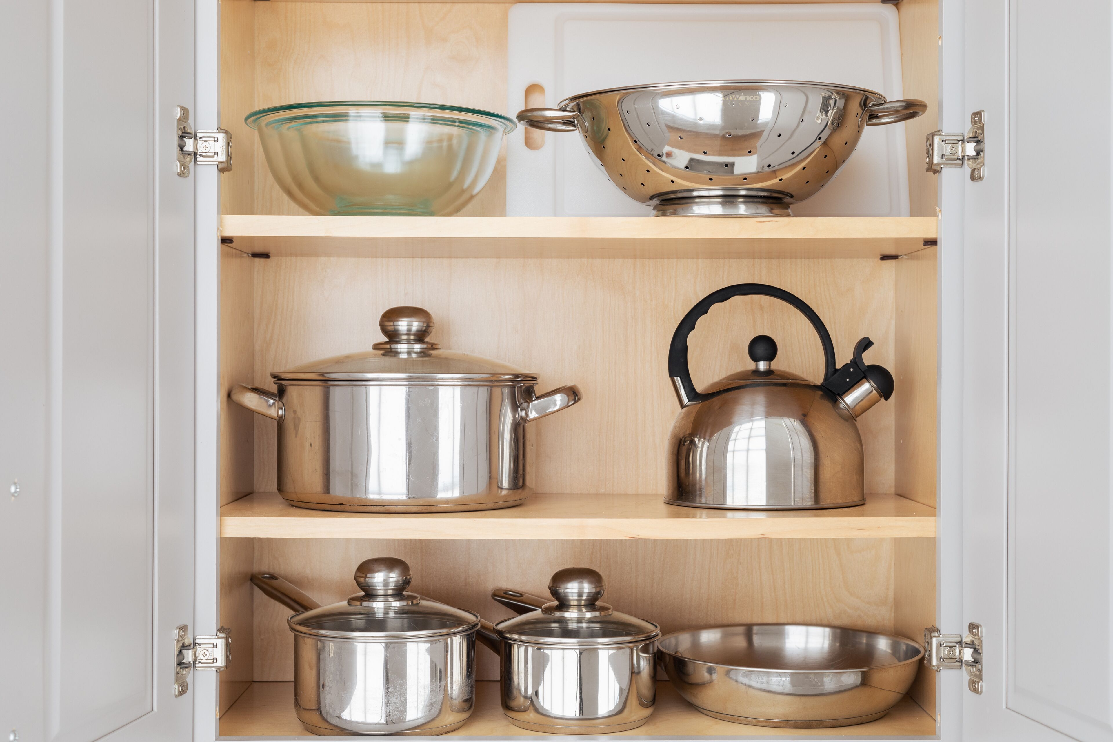 A kitchen cabinet with stainless steel cookware, including pots, a kettle, and a colander.