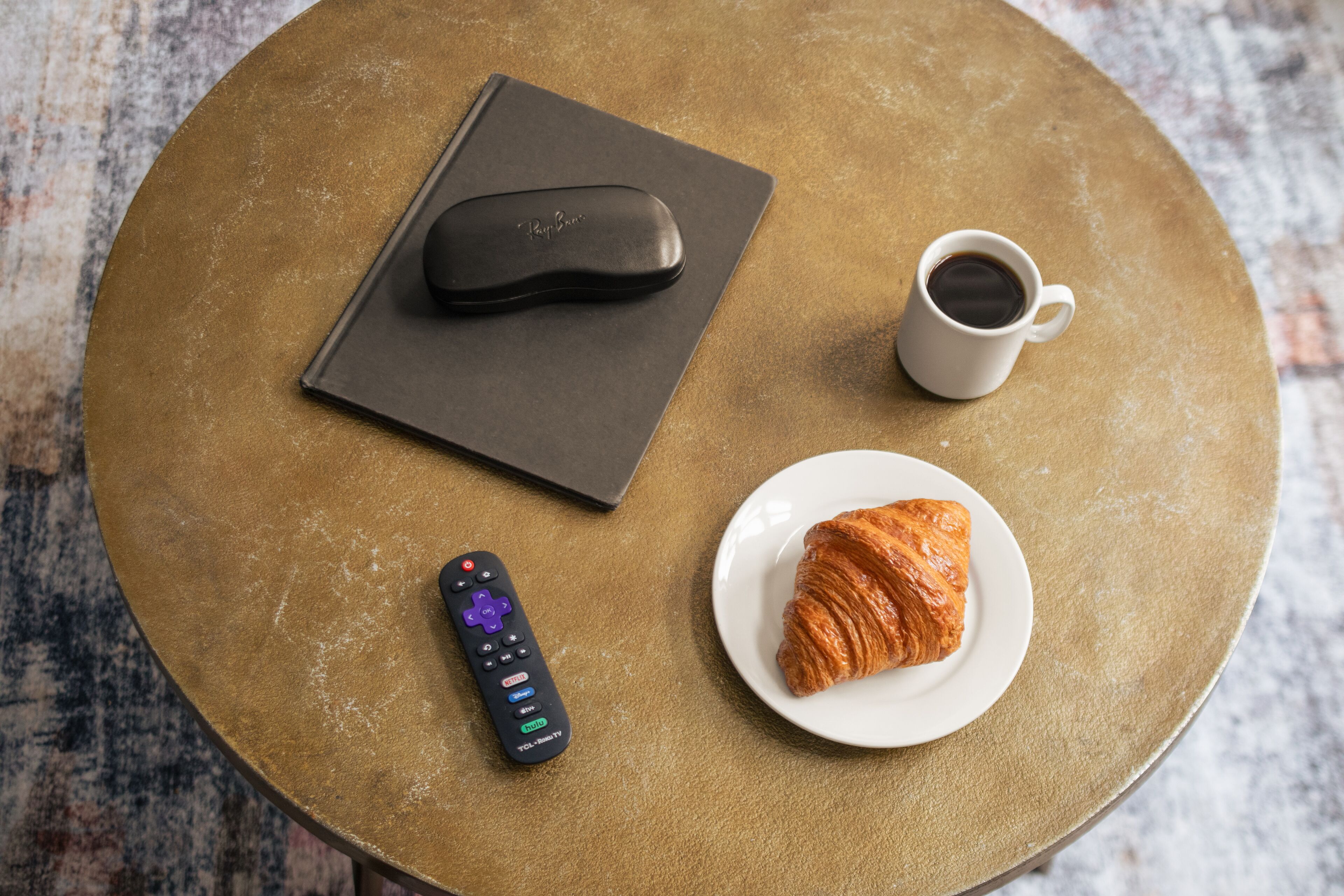 A round table with a remote control, a cup of coffee, a croissant, and a book.