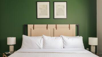 A neatly made bed with white linens, a headboard with a brown panel, and two framed artworks on the green wall above.