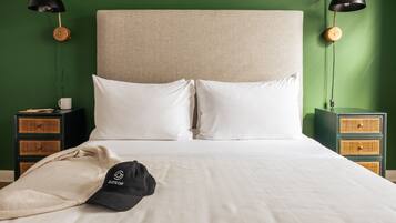 A neatly made bed with white linens, a single black cap on the bed, and two bedside tables with black lamps and wicker storage drawers against a green wall.