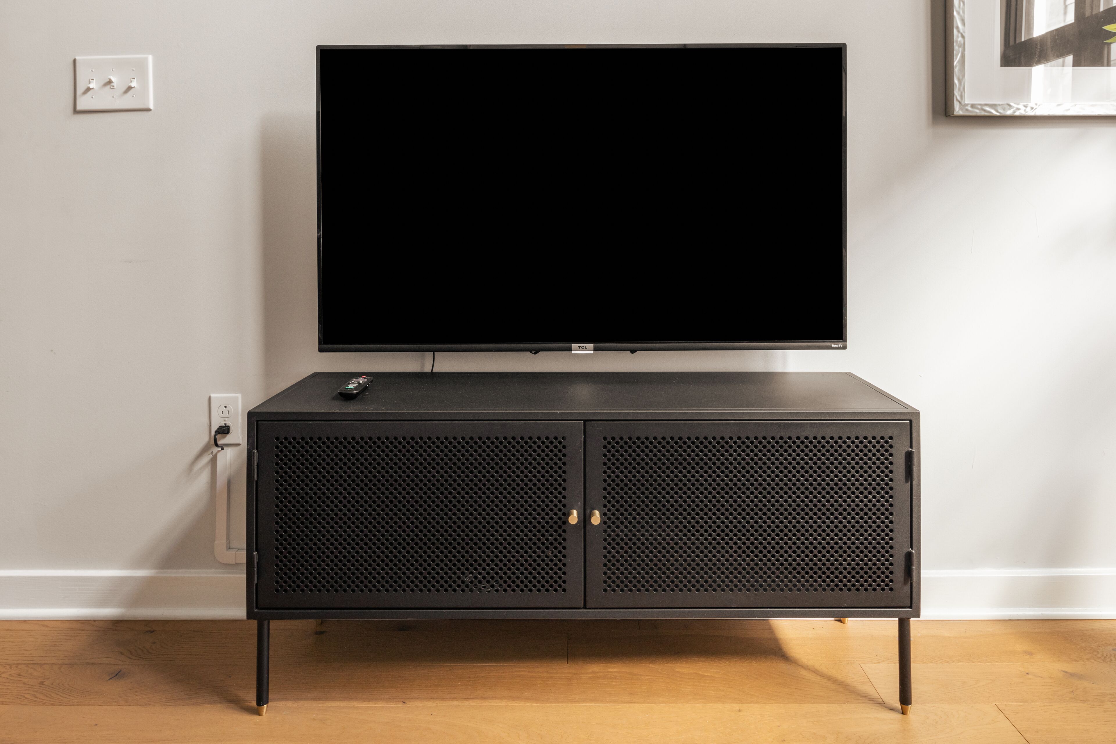 A black TV stand with a flat-screen TV on top, a power outlet, and a framed picture on the wall.