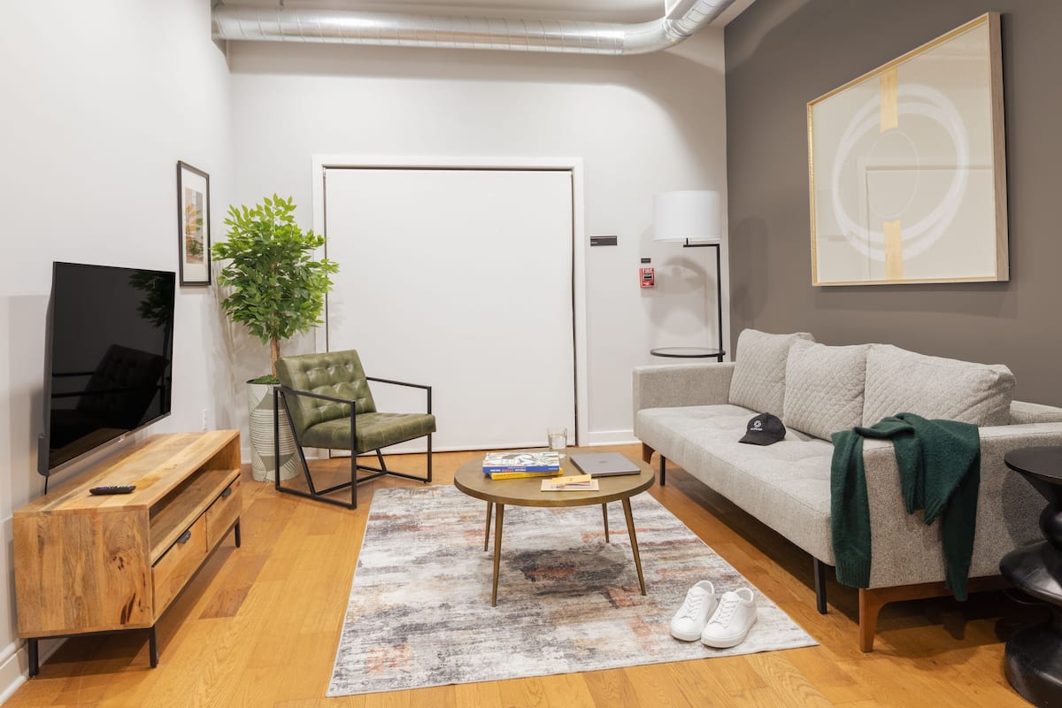 Ein modernes Wohnzimmer mit Flachbildfernseher, einem grünen Sessel, einem grauen Sofa, einem Couchtisch und einem großen abstrakten Gemälde an der Wand.