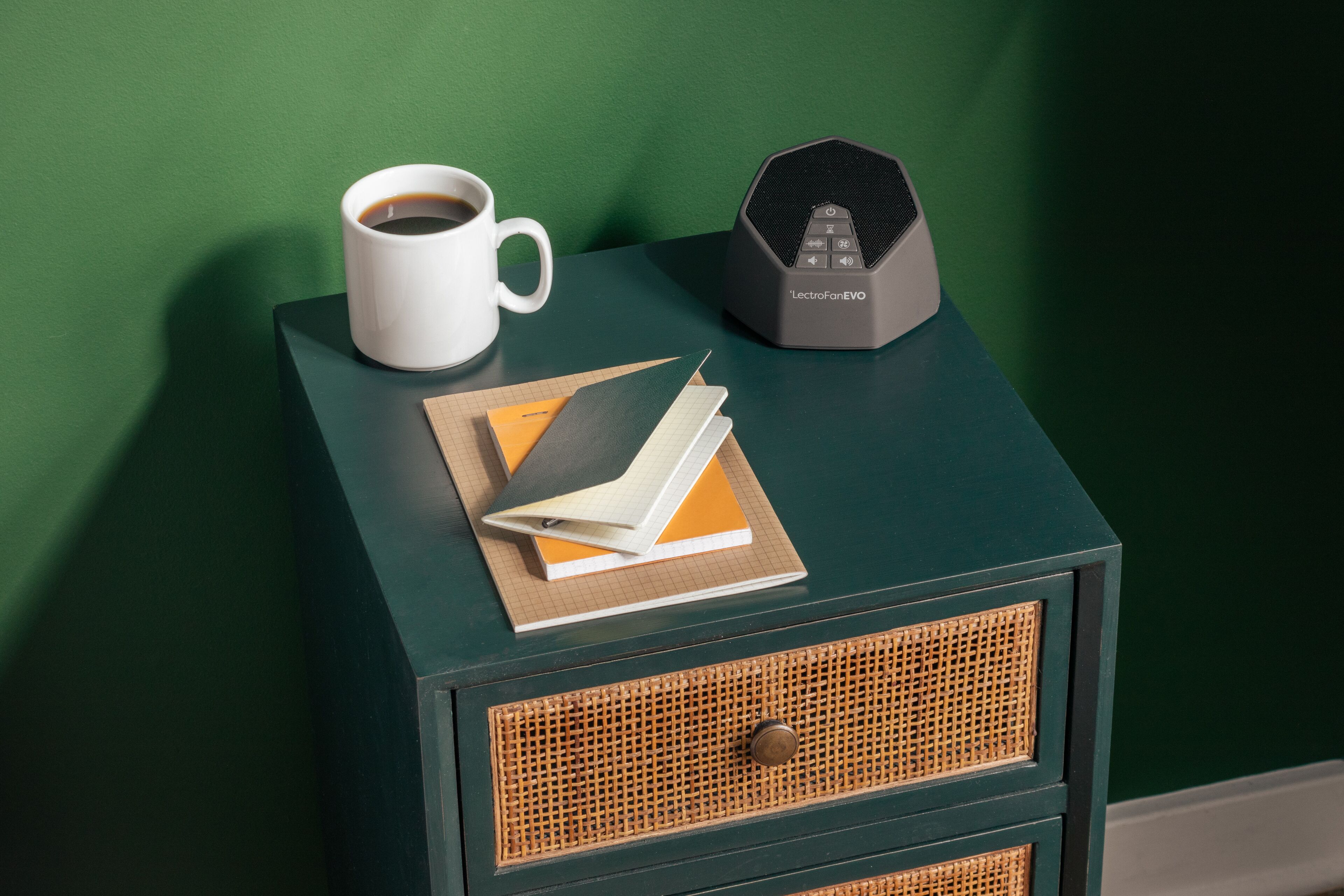 Una mesita de noche pequeña con una taza blanca, un altavoz negro y un cuaderno.