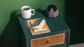 Una mesita de noche pequeña con una taza blanca, un altavoz negro y un cuaderno.