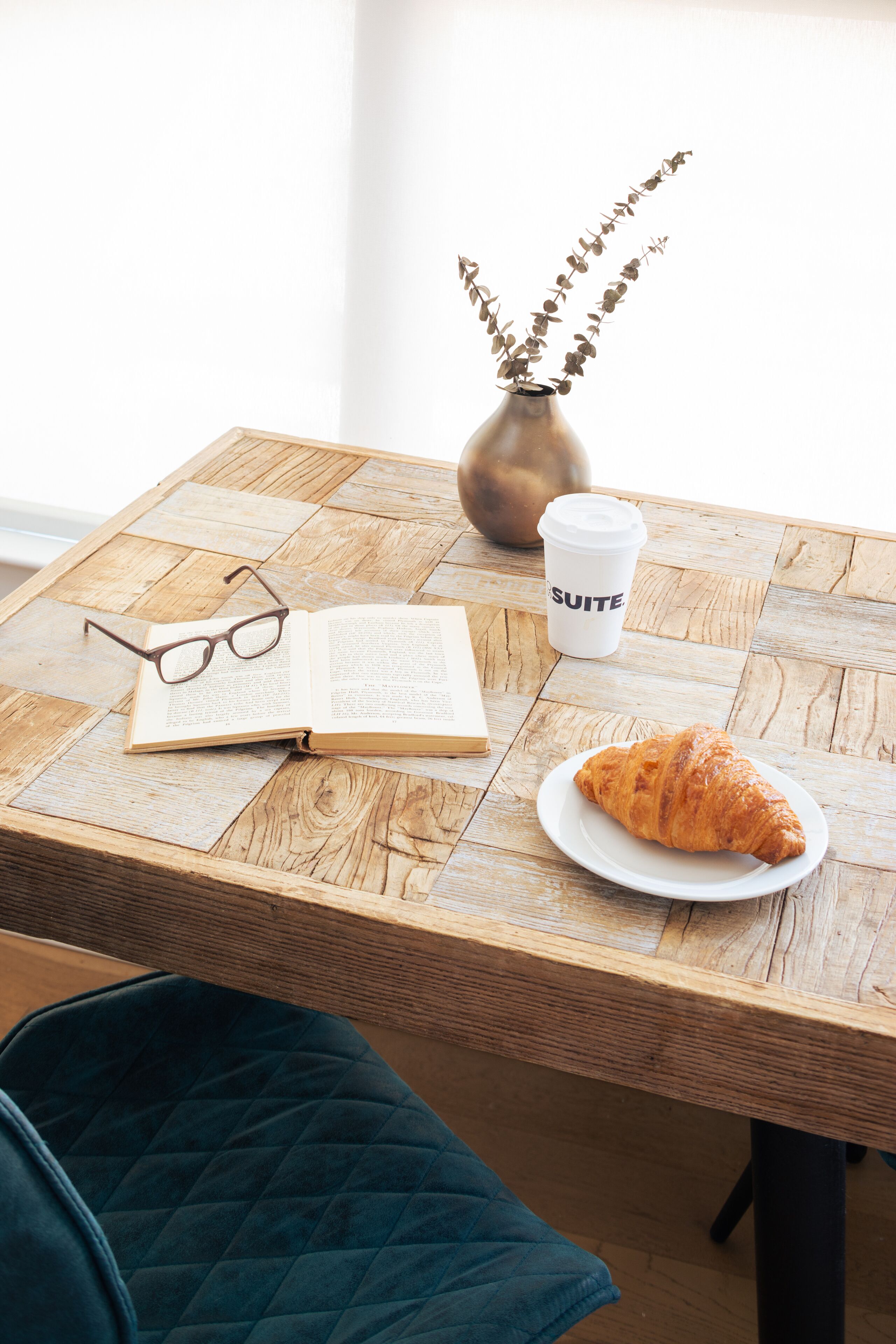 Een houten tafel met een boek, een bril, een koffiekop, een vaas met gedroogde bloemen en een croissant op een bord.