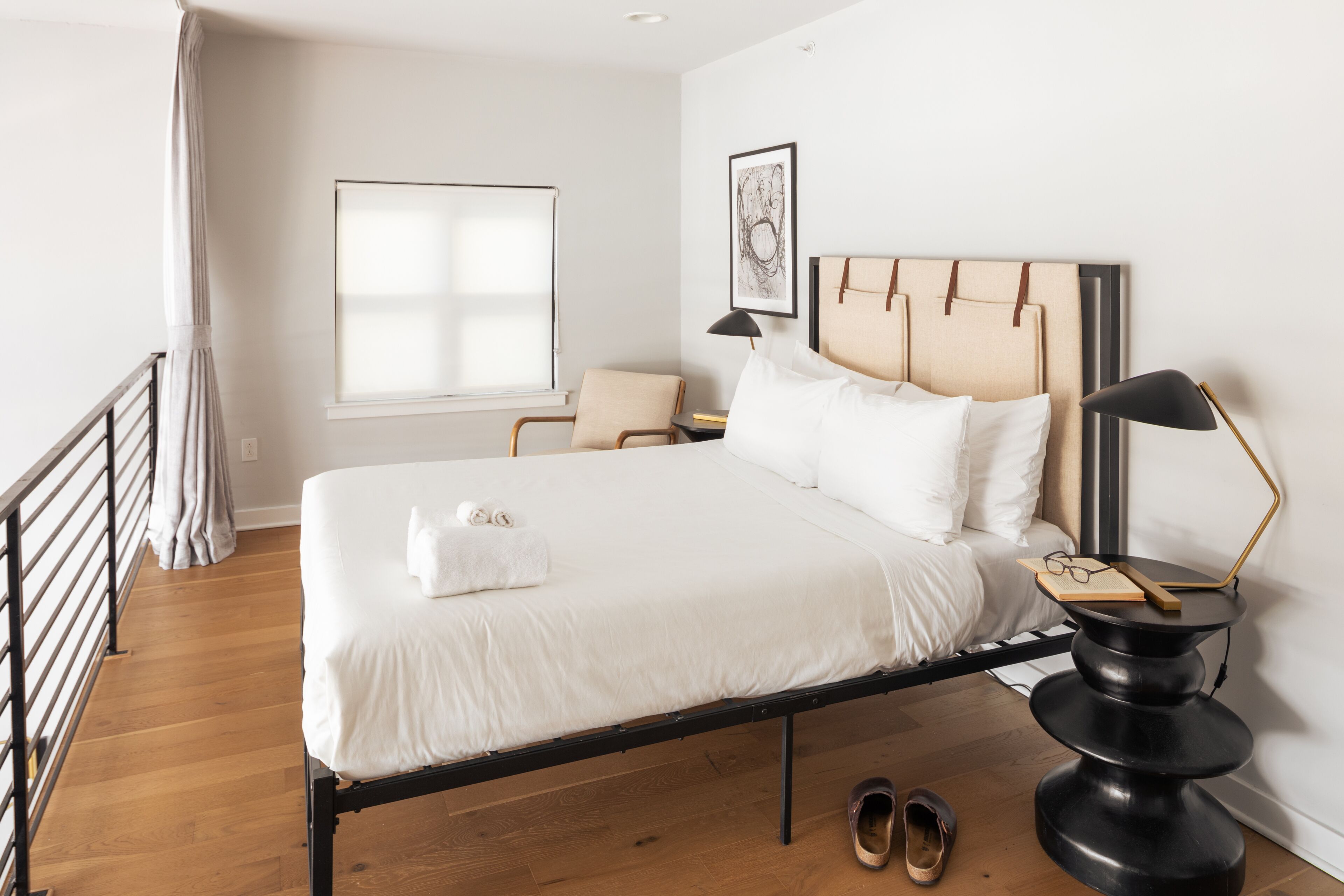A neatly made bed with white linens, a bedside table with a lamp, and a pair of shoes on the floor.