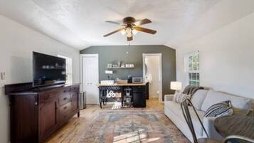 A compact living space with a ceiling fan, a flat-screen TV, a dining area with a table and chairs, and a sofa with cushions.