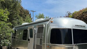 Een zilveren Airstream caravan op een grindpad, met een blauwe prullenbak in de buurt.