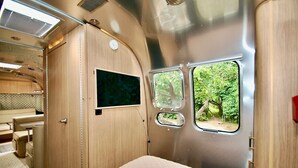 Een compact interieur van een camper met een bed, een kleine televisie en een raam met uitzicht op begroeiing.