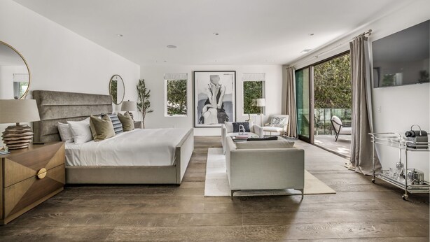 A modern bedroom with a large bed, a sofa, and a TV. There is a window with curtains and a view of greenery outside.