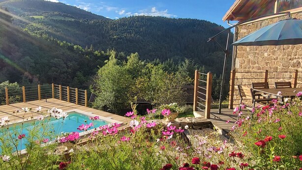 Un área de piscina con un deck de madera, rodeada de flores y un edificio de piedra también con deck y bancos.