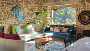 Una sala de estar acogedora con pared de piedra, un sofá azul, un sofá blanco, un televisor y un ventanal con vistas a árboles.