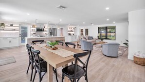 A modern kitchen with white cabinetry, a dining area with a wooden table and black chairs, and a living area with a grey sofa and a large window.