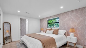 A bedroom with a large bed, a nightstand, a mirror, and a window with a view of greenery.