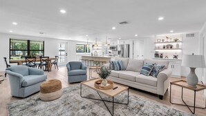 A modern living room with a sofa, armchairs, and a coffee table. There is a dining area with a table and chairs. The kitchen is visible with appliances and cabinets.