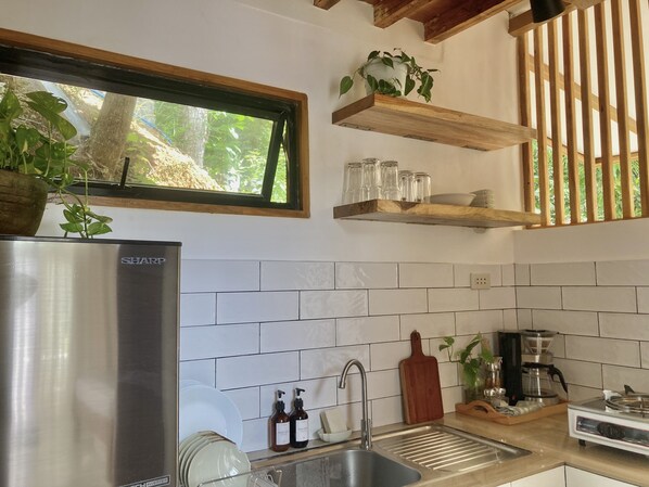 A modern kitchen with a stainless steel refrigerator, white tiled backsplash, wooden shelves, and a window with a view of greenery.