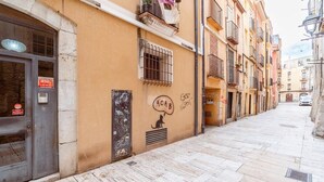Uma rua estreita com edifícios em ambos os lados, uma loja fechada com porta de metal e um muro com grafites e desenho de um gato.
