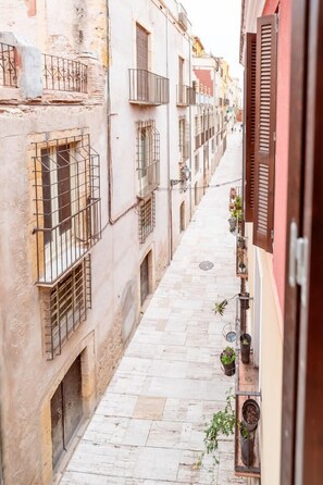 A narrow alleyway with buildings on either side, featuring balconies with metal railings and potted plants.