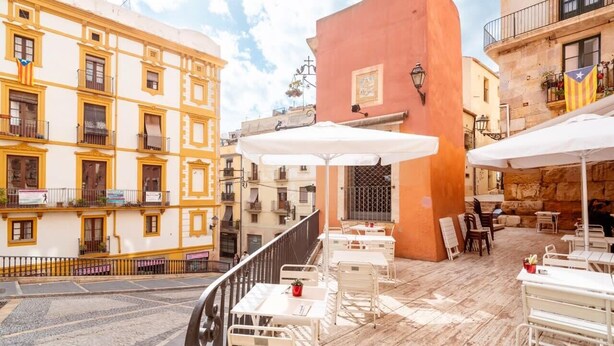 Um terraço com mesas ao ar livre, um edifício vermelho e edifícios amarelos ao fundo.