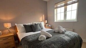 A neatly made bed with a grey blanket and pillows, a bedside lamp, and a window with blinds. - Nollaig  Beachside Bliss - Pass the Keys (Rye)