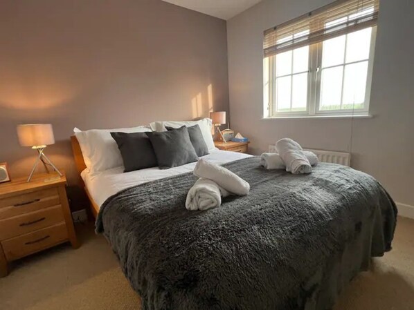 A neatly made bed with a grey blanket and pillows, a bedside lamp, and a window with blinds.