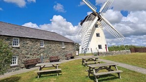 A traditional windmill with a stone building adjacent to it, featuring picnic tables and benches in the foreground. - Self Contained Apartment at Country Pub Restaurant - Pass the Keys (Llanddona)