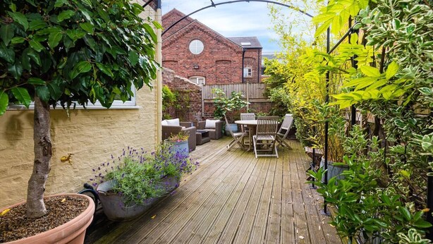 A wooden deck with outdoor furniture, surrounded by potted plants and a brick building in the background.