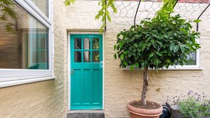 A turquoise door with a small tree in a pot beside it, a window, and a potted plant with purple flowers on the porch.
