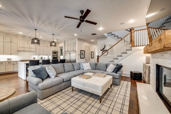 A spacious living room with a large grey sectional sofa, a fireplace, and a staircase with wooden railings. - Modern Serenity: 3 Beds of Calm & Style (Dallas)