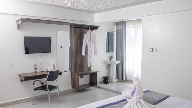A hotel room with a bed, a desk, a chair, a TV, a mirror, a sink, and a robe hanging on a rack. - WAFFLES SUITES (Nakuru)