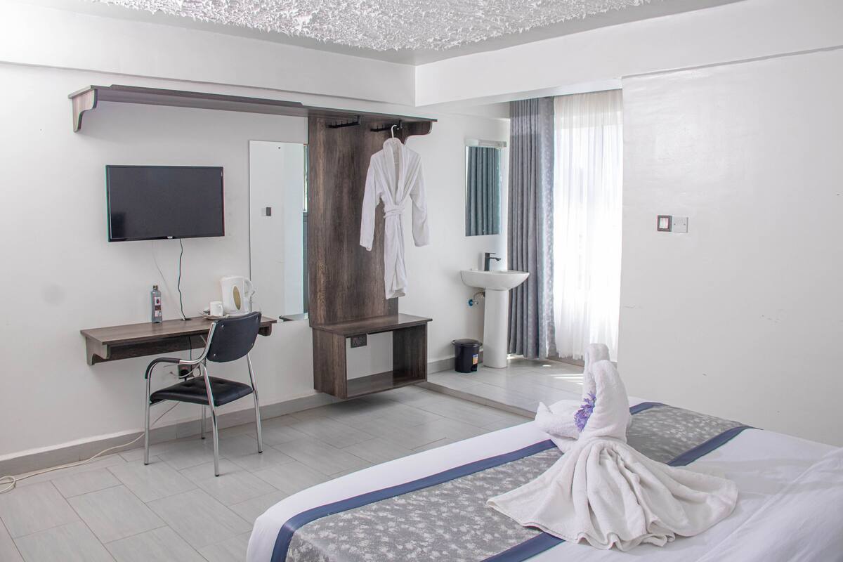 Habitación de hotel con cama, escritorio, silla, televisor, espejo, lavabo y bata colgada en un perchero.