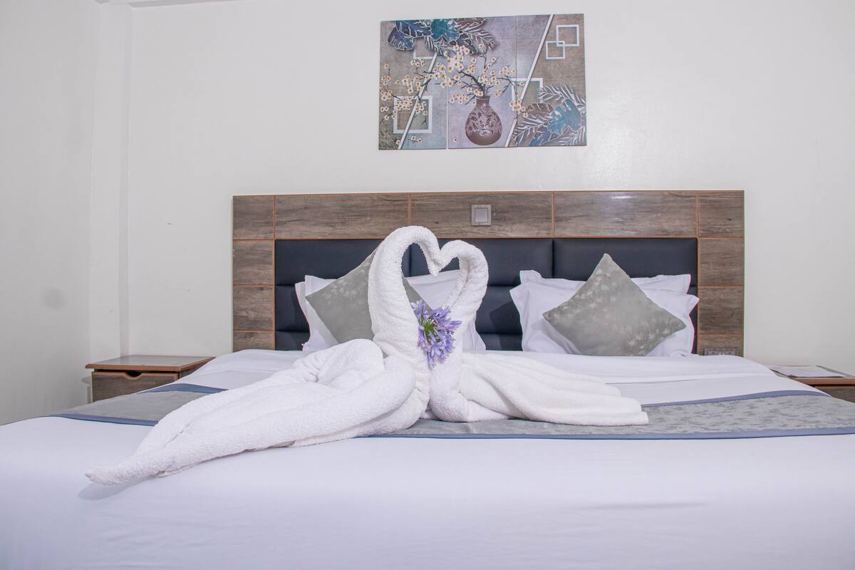 Una cama bien hecha con toallas dispuestas en forma de corazón, un cabecero de madera y un cuadro en la pared.
