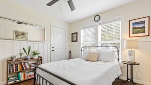 A bedroom with a bed, a bookshelf, a clock, and a window with blinds. - Mary’s Mid-Century Modern House- 3-Bed Haven (Fort Worth)