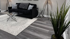 Uma sala de estar moderna com um sofá preto, uma mesa de centro de vidro e um tapete branco grande. Há duas luminárias de chão e um vaso de plantas.