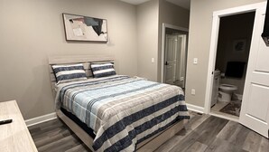 Quarto com cama, mesa de cabeceira, porta e banheiro visível por uma porta aberta.