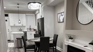 Uma cozinha moderna com armários brancos, uma sala de jantar com mesa e cadeiras redondas, e uma lareira com fogo vermelho.