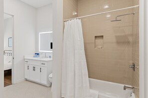 A modern bathroom with a white vanity, a large mirror, a shower with a beige tiled wall, and a white bathtub. - The Corbin 1307: Princeton 1-Bedroom Elegance (Fort Worth)