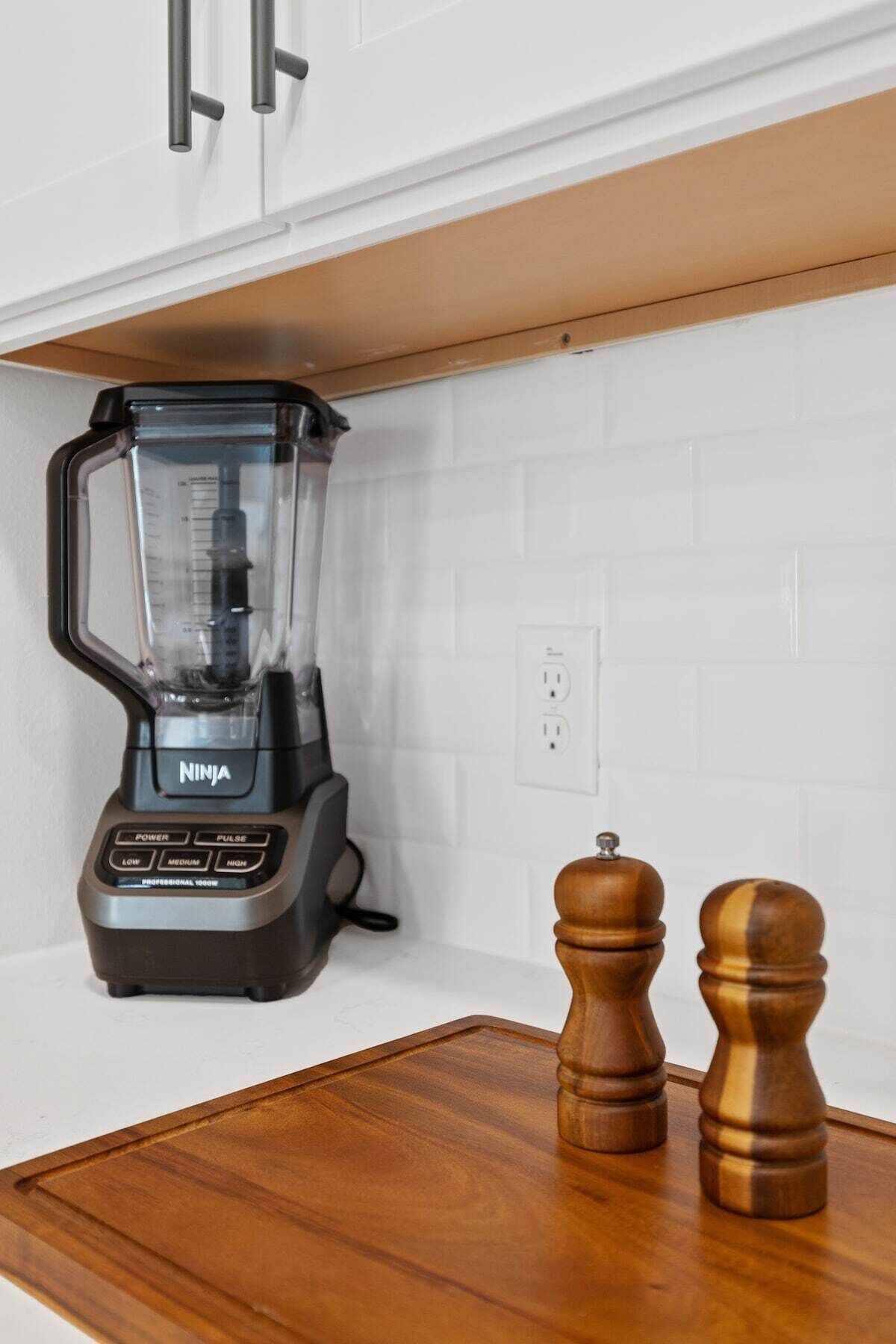 A kitchen with a Ninja blender on a wooden countertop, a white tiled backsplash, and wooden salt and pepper shakers.