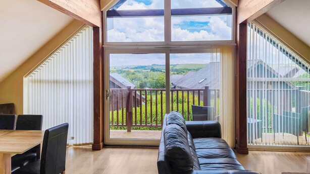 A spacious room with a wooden ceiling, a large sliding glass door leading to an outdoor balcony, a black leather sofa, and a wooden dining table with chairs.