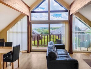 A spacious room with a wooden ceiling, a large sliding glass door leading to an outdoor balcony, a black leather sofa, and a wooden dining table with chairs.