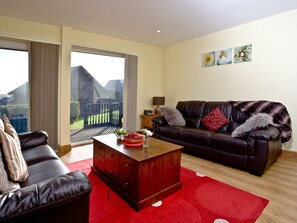 A living room with a leather sofa, a wooden coffee table, and a red rug. There is a sliding glass door leading to a balcony. - Beech Lodge - Retallack Resort (St Columb)