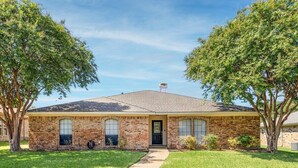 A two-story brick house with a front entrance, a covered porch, and a lawn with two trees. - Cozy Nest-3-Bedroom Charm In Plano (Plano)