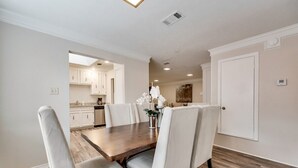 A dining area with a wooden table and white upholstered chairs, a vase with flowers, and a kitchen in the background.