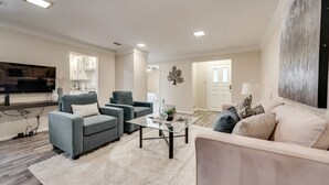 A modern living room with a grey sofa, a beige sofa, and a glass coffee table. There is a flat-screen TV mounted on the wall, and a decorative tree on the wall.