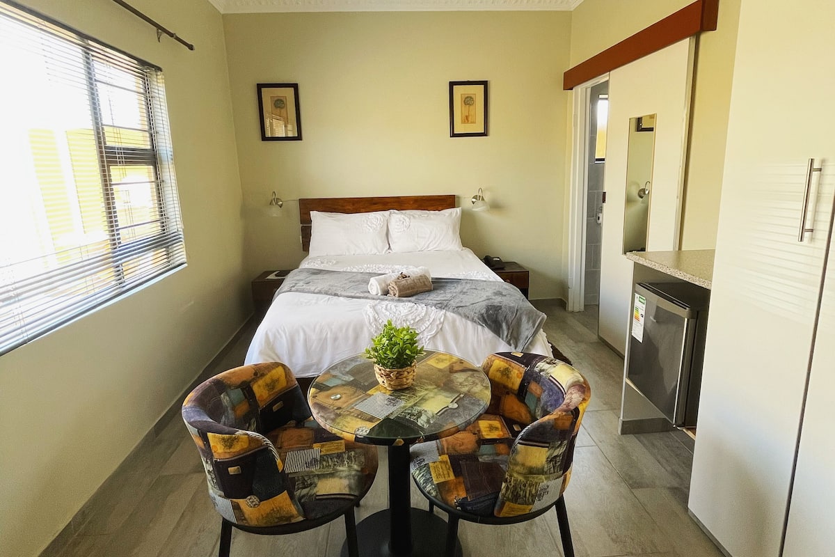 Habitación de hotel con cama, una pequeña mesa con sillas, una planta en maceta y una kitchenette.